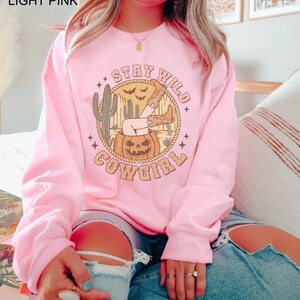 May include: A light pink sweatshirt featuring a graphic print of a woman's leg sticking out of a jack-o-lantern. The graphic also includes the text "Stay Wild Cowgirl" and a cactus.