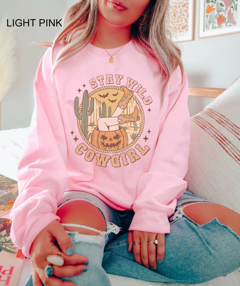 May include: A light pink sweatshirt featuring a graphic print of a woman's leg sticking out of a jack-o-lantern. The graphic also includes the text "Stay Wild Cowgirl" and a cactus.