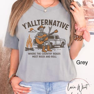 May include: Grey t-shirt with the text "Y'ALLTERNATIVE" above an illustration of a skeleton playing a guitar next to a truck and campfire. The text "WHERE THE COUNTRY ROADS MEET ROCK AND ROLL" is below the illustration.
