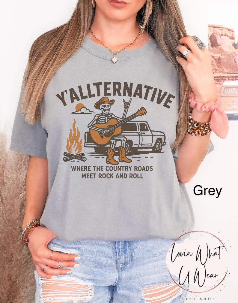 May include: Grey t-shirt with the text "Y'ALLTERNATIVE" above an illustration of a skeleton playing a guitar next to a truck and campfire. The text "WHERE THE COUNTRY ROADS MEET ROCK AND ROLL" is below the illustration.