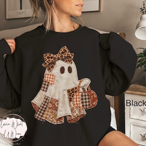 Op de afbeelding: Een zwart sweatshirt met ronde hals en een centrale afbeelding van een geest versierd met een strik met luipaardprint. De geest is samengesteld uit verschillende stofachtige patches in oranje, bruine en witte patronen en houdt een kleine pompoen vast.