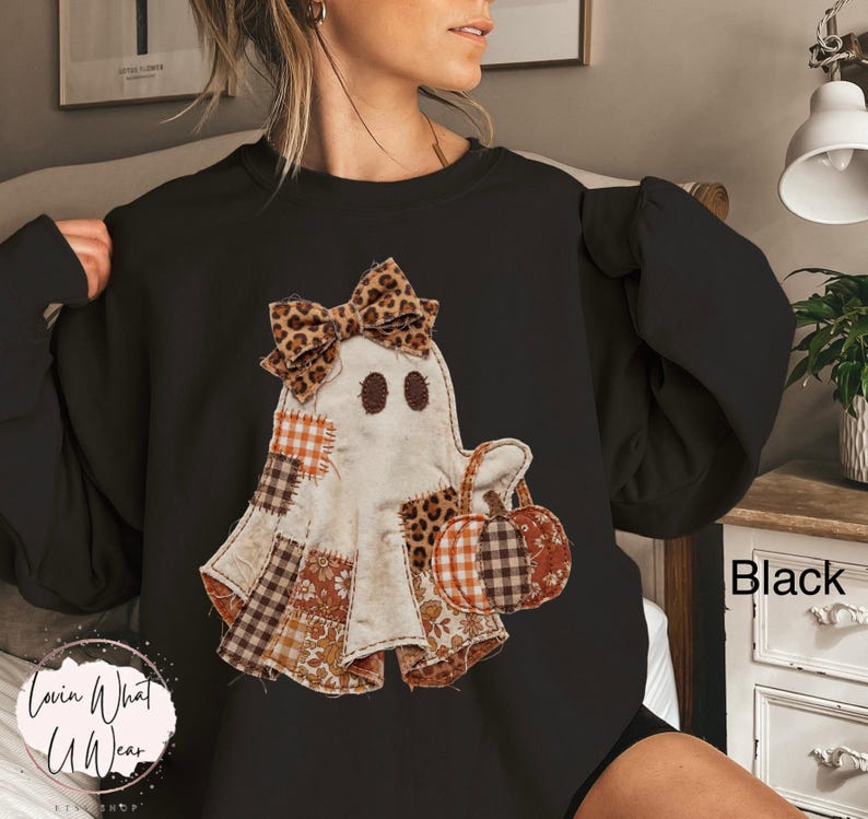 Op de afbeelding: Een zwart sweatshirt met ronde hals en een centrale afbeelding van een geest versierd met een strik met luipaardprint. De geest is samengesteld uit verschillende stofachtige patches in oranje, bruine en witte patronen en houdt een kleine pompoen vast.
