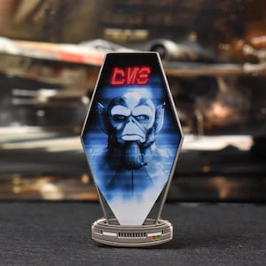 Könnte beinhalten: Ein blau-weißes holografisches Display mit einer Figur und dem roten Text "DWE". Das Display steht auf einem silbernen Sockel mit roten und grünen Lichtern, mit einem verschwommenen Bild eines Raumschiffs im Hintergrund.