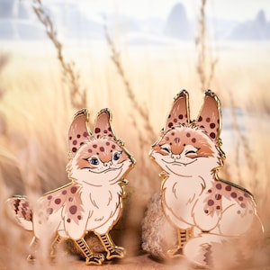 Könnte beinhalten: Zwei goldfarbene Emaille-Pins mit einem Cartoon-Wüstenkatze mit braunen Flecken. Die Katze links steht mit dem Schwanz hinter dem Rücken eingerollt. Die Katze rechts sitzt mit dem Schwanz um die Beine gerollt. Beide Katzen haben ein weißes Gesicht und eine braune Nase.