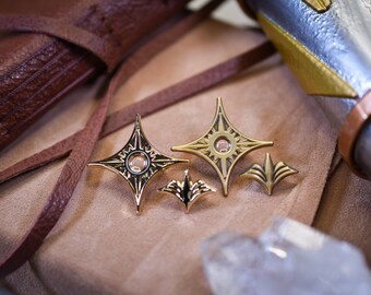 May include: Three gold-colored metal pins with a starburst design. The pins have a raised center with a small circle in the middle. The pins are on a brown leather surface.
