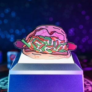 Op de afbeelding: Een roze en witte emaille pin van een cartoon hotdog met toppings, op een witte displaystandaard. De pin is een nieuwigheidsvoedsel.