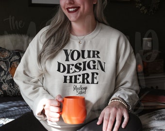May include: A person wearing a light beige sweatshirt with the text "YOUR DESIGN HERE" and the words "Mockup Sisters" in smaller font. The person is holding an orange mug.