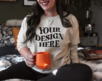 May include: A person wearing a beige sweatshirt with the text "YOUR DESIGN HERE" in black. The person is holding an orange mug.