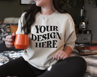 May include: A person wearing a beige sweatshirt with the text "YOUR DESIGN HERE" in black. The person is holding an orange mug and sitting on a bed.