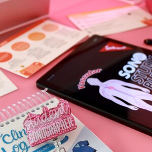 May include: A pink desk setup featuring a tablet displaying a medical diagram, a student sonographer badge, and a spiral-bound clinical log. The badge is glittery and reads "Student Sonographer". Other items include color charts and a stapler.