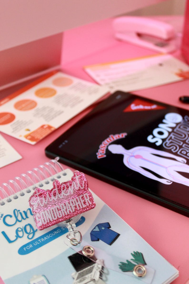 May include: A pink desk setup featuring a tablet displaying a medical diagram, a student sonographer badge, and a spiral-bound clinical log. The badge is glittery and reads "Student Sonographer". Other items include color charts and a stapler.