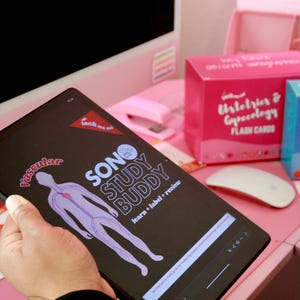 May include: A tablet displaying the "SONO STUDY BUDDY" app with a vascular diagram. Pink and blue flashcard boxes for "Histology & Gynecology" and "Ultrasound Physics" are visible, along with a white mouse, on a pink desk.