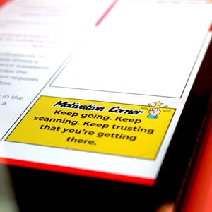 May include: A close-up of a document with a yellow sticky note in the corner. The note reads "Motivation Corner" and includes motivational text. The document has a red border and is on a black surface.