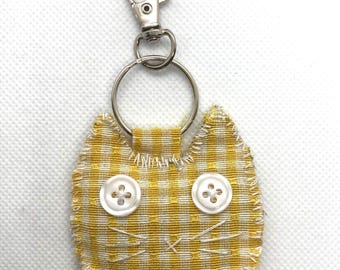 May include: A yellow and white gingham cat-shaped keychain. The cat's face is detailed with two white buttons for eyes and stitched whiskers. It has a silver-colored metal clasp and keyring.