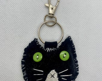 May include: A black cat-shaped keychain with green button eyes and white stitched whiskers and mouth. The cat is made of felt and has a silver-colored clasp and keyring.