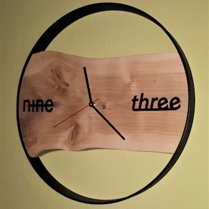 Könnte beinhalten: Ein hölzernes Zifferblatt mit schwarzen Zeigern und schwarzen Zahlen für neun und drei, in einem schwarzen runden Rahmen.