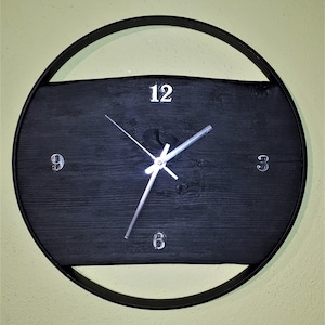 Könnte beinhalten: Eine schwarze Uhr mit silbernen Zeigern und römischen Ziffern auf einem schwarzen Holzhintergrund. Die Uhr ist in einen schwarzen Metallkreis gefasst.