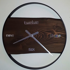 Könnte beinhalten: Ein dunkelbraunes Holzuhrblatt mit silbernen Metallziffern und Zeigern. Das Uhrblatt ist von einem schwarzen Metallring umgeben. Die Zahlen auf dem Uhrblatt sind zwölf, neun, sechs und drei.