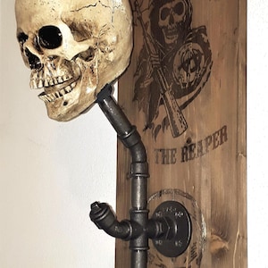 Peut inclure: Un porte-manteau mural en métal noir avec un crâne sur le dessus. Le porte-manteau est monté sur une planche de bois avec les mots "Sons of Anarchy" et "The Reaper" imprimés dessus.