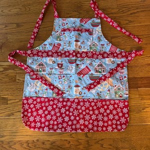 Fun festive Christmas apron, decorated with gingerbread houses, gingerbread men, snow man, and other decor . pocket of red snowflake accent.