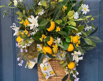 May include: A decorative basket overflowing with faux lemons, white and yellow flowers, and green leaves. A lemon-themed ribbon adorns the basket, which is hanging on a blue door. The arrangement is a vibrant and cheerful display.