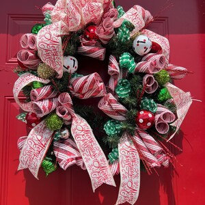 May include: A festive Christmas wreath with red and white striped ribbon, green and red ornaments, and sprigs of evergreen. The wreath is decorated with white ribbon printed with "Merry Christmas". The wreath is displayed on a red door.