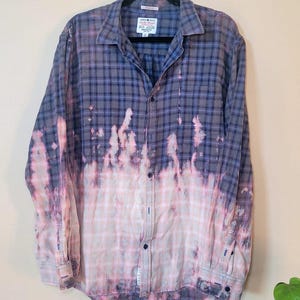 May include: A blue and white plaid flannel shirt with a bleach-dyed effect. The shirt has a button-up front and long sleeves.