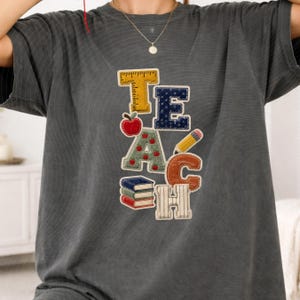 May include: A charcoal gray t-shirt featuring the word TEACH spelled out with school-themed graphics. The T is a yellow ruler, the E is navy blue with white dots, the A is green with red apples, the C is a pencil, and the H is books.