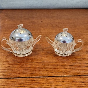 May include: Two clear glass salt and pepper shakers shaped like teapots with silver lids. The shakers are on a wooden surface.
