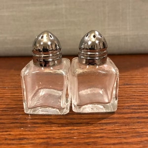 May include: A set of two clear glass salt and pepper shakers with silver tops. The shakers are square shaped and have a small hole in the top for dispensing salt and pepper.