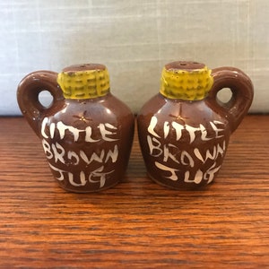 May include: A pair of brown ceramic salt and pepper shakers shaped like jugs. The shakers are decorated with yellow corn husks and white lettering that reads "Little Brown Jug".