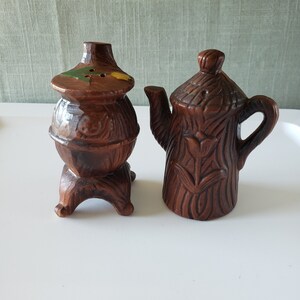 May include: Two ceramic salt and pepper shakers. The salt shaker is shaped like a wood-burning stove with a brown glaze and green and yellow accents. The pepper shaker is shaped like a teapot with a brown glaze and a floral design.