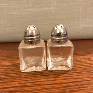 May include: Two clear glass salt and pepper shakers with silver tops. The shakers are square shaped and have a small hole in the top for dispensing salt and pepper.