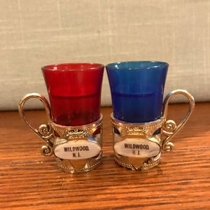 May include: Two small shot glasses with silver holders. The glasses are red and blue. The holders have a silver band with the words "WILDWOOD N.J." on them.