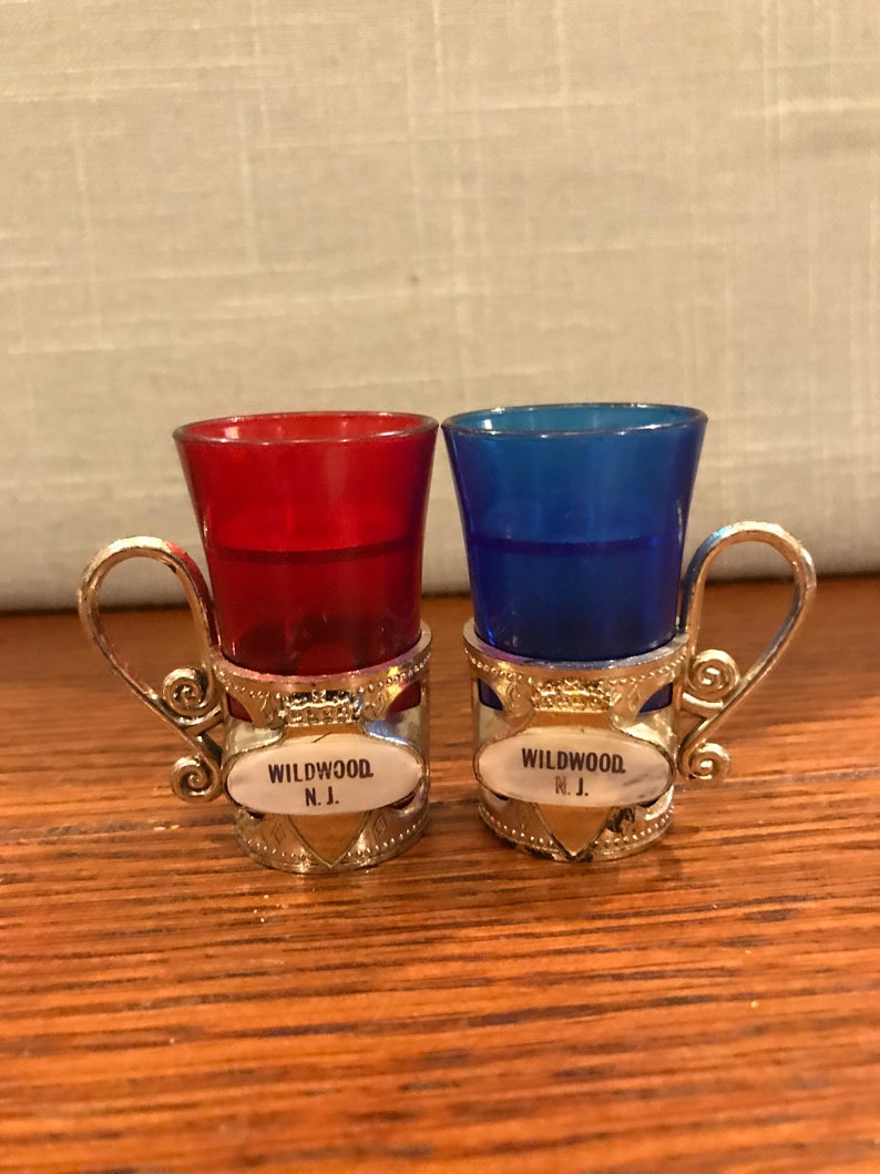 May include: Two small shot glasses with silver holders. The glasses are red and blue. The holders have a silver band with the words "WILDWOOD N.J." on them.