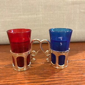 May include: A set of two shot glasses, one red and one blue, with silver metal holders. The holders are connected by a silver metal handle.