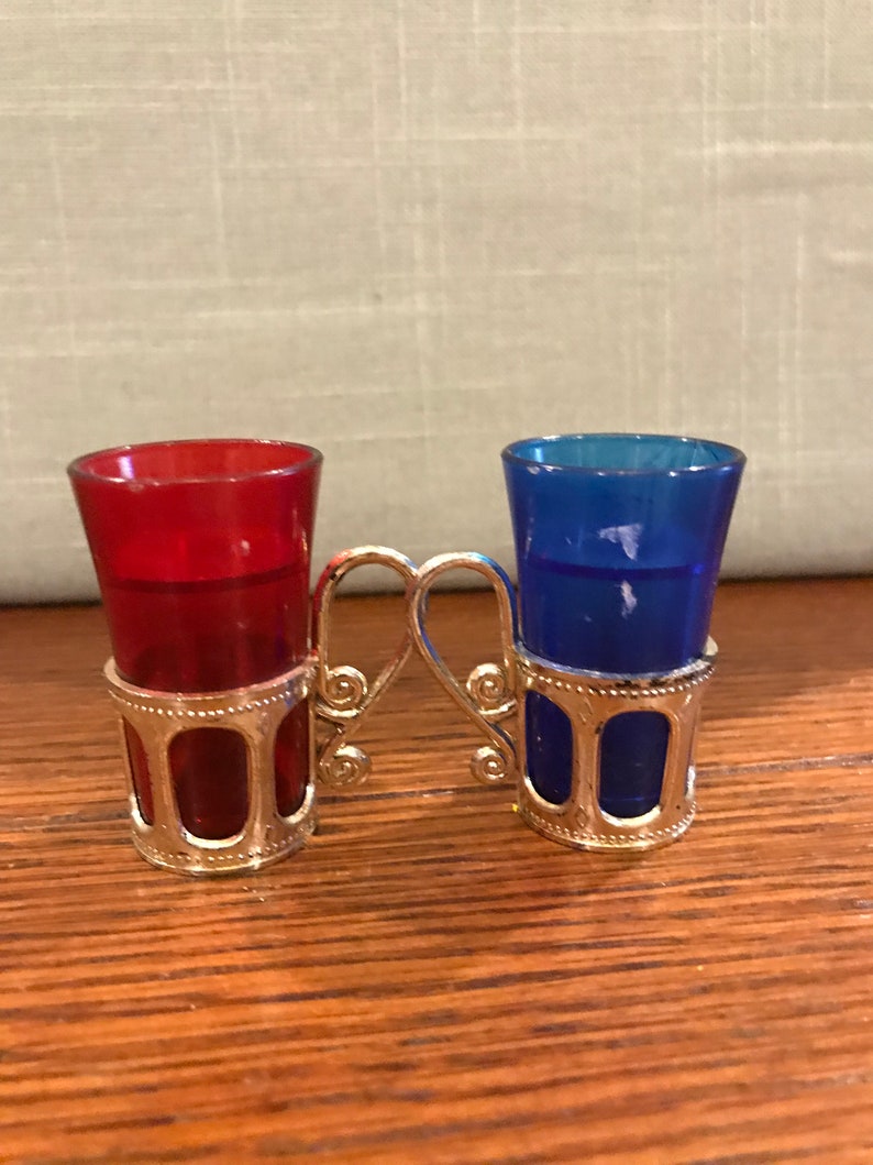 May include: A set of two shot glasses, one red and one blue, with silver metal holders. The holders are connected by a silver metal handle.
