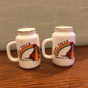 May include: A pair of white ceramic salt and pepper shakers shaped like mugs. They feature a gold rim and a colorful image of Ruby Falls, Lookout Mountain, Tennessee.
