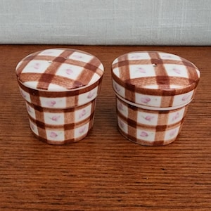 May include: A set of two ceramic salt and pepper shakers. The shakers are shaped like small pots and are decorated with a brown and white checkered pattern with pink flowers.