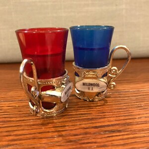 May include: Two small shot glasses with silver holders. The glasses are red and blue. The holders have a silver handle and a small oval tag that says "WILDWOOD, N.J."
