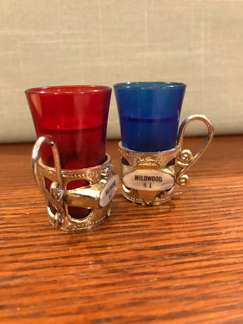 May include: Two small shot glasses with silver holders. The glasses are red and blue. The holders have a silver handle and a small oval tag that says "WILDWOOD, N.J."