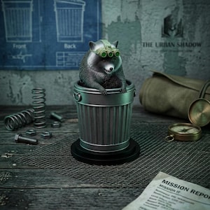 May include: A figurine of a raccoon wearing night vision goggles, sitting inside a miniature trash can. The scene includes a blueprint labeled "trash tech," a compass, and a "Mission Report" document, creating a playful, urban theme.