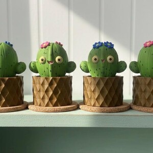 May include: Four green cactus-shaped planters with smiling faces and small flower accents. Each cactus sits in a brown geometric pot on a cork base. The planters are arranged on a light green shelf.