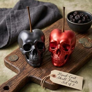 May include: Two skull-shaped decorative objects, one metallic gray and one metallic red, with brown stems, resembling cherries. They sit on a wooden cutting board with a tag that reads "Hand-Cast Skull Fruits - Set of 2."