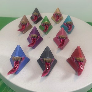 May include: A set of 10 pyramid-shaped dice with a tongue sticking out. The dice are painted in various colors, including red, blue, green, purple, gold, and black.