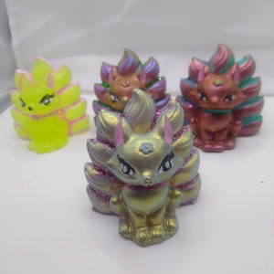 May include: Four colorful kitsune figurines with iridescent paint and glitter. Each kitsune has a different color scheme, including yellow, purple, green, and red.