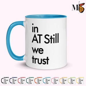 May include: White ceramic mug with a blue interior and handle. The mug features the text "in AT Still we trust" in black, centered on the front. A butterfly logo is visible in the upper right corner. Several mugs with different colored handles are shown below.