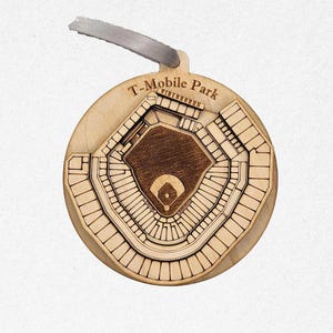 May include: A wooden ornament in the shape of T-Mobile Park, a baseball stadium. The ornament is round with a detailed laser-cut design of the stadium's layout. It has a light brown color with a gray ribbon for hanging.