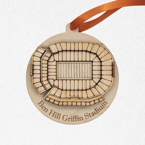 May include: A wooden ornament shaped like a stadium, with the text "Ben Hill Griffin Stadium" engraved on the bottom. The ornament has a light brown color and an orange ribbon for hanging.