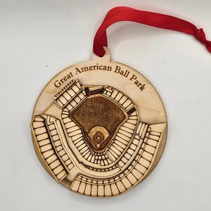 May include: A wooden ornament featuring a detailed laser-cut design of a baseball stadium. The ornament is round with the text "Great American Ball Park" engraved on the top. A red ribbon is attached for hanging.